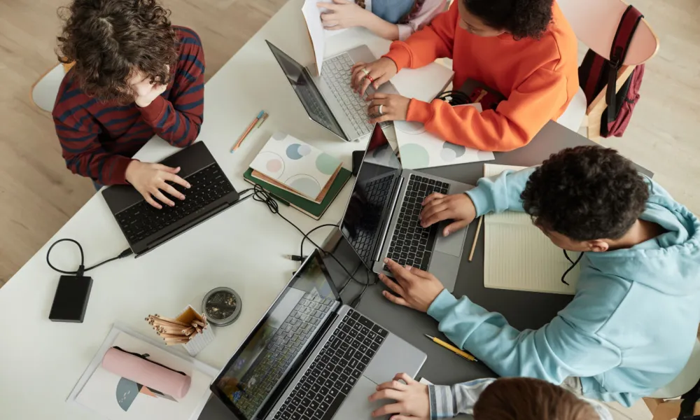 Top view at diverse group of teen school children using computers in classroom studying together at table for Governor's School of Computer Science.