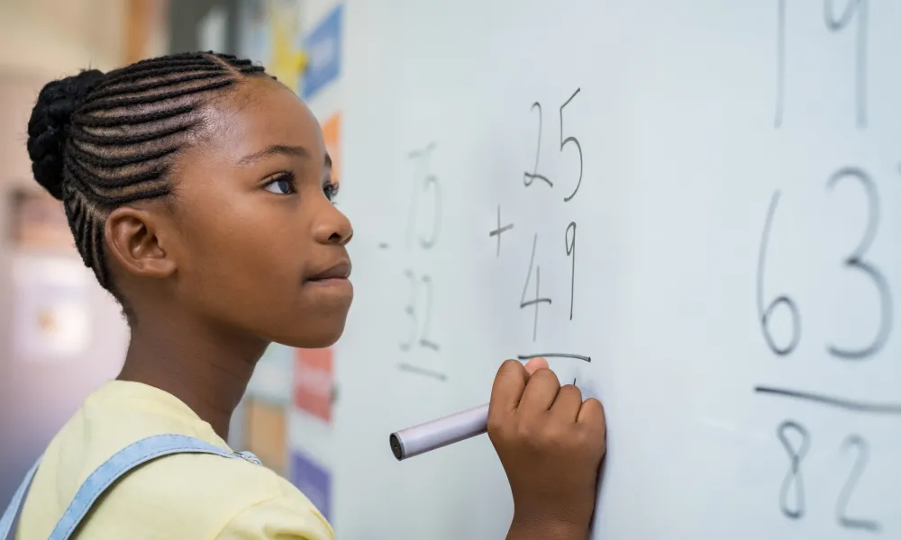 A young female holds a marker while staring with determination at an addition problem on the whiteboard.