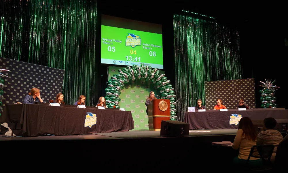 Students sitting at tables and a woman standing behind a podium.