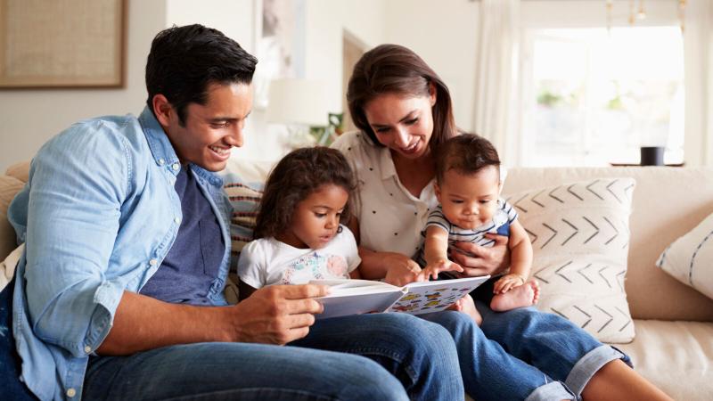 Young Hispanic family of four sitting on the sofa reading a book together in their living room