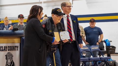 Older veteran receiving diploma at event.  Two people standing to either side to congratulate him, crowd behind