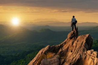 Young man holding business bag on top of peak mountain and looking into the sunset.