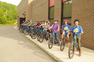 Several young school children on bicycles on sidewalk.  Large brick building behind