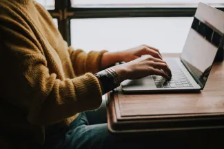 A female in a cozy sweater on a fall day types on a laptop keyboard.