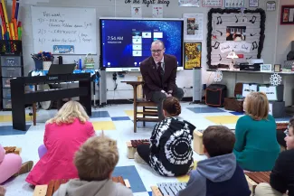 Teacher sitting on a chair in front of students in a classroom.